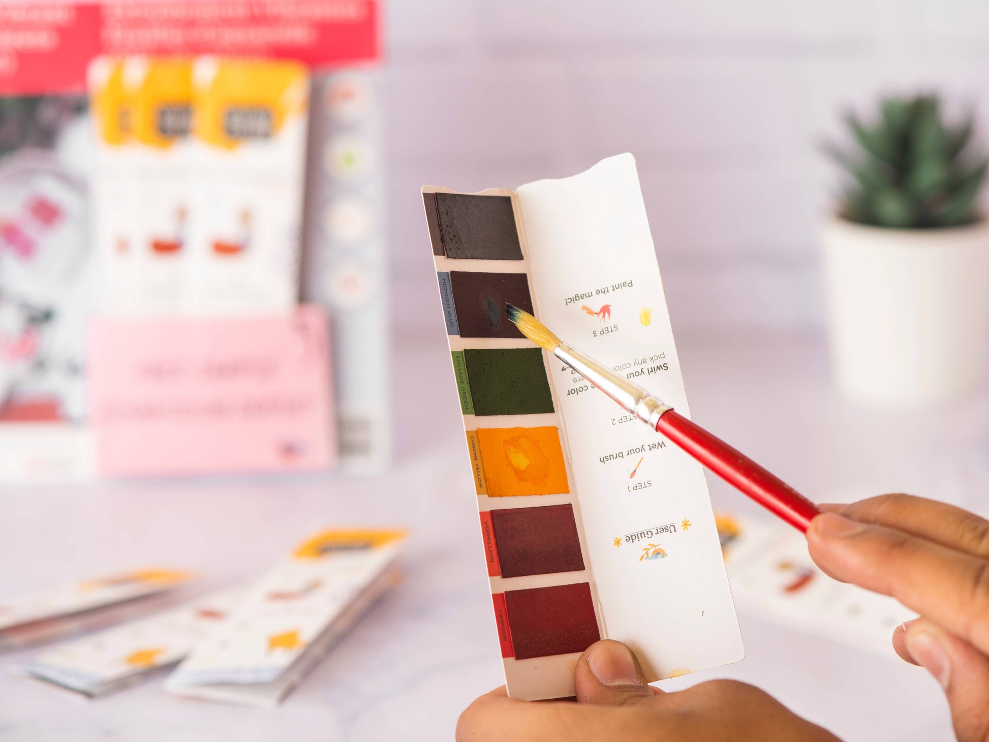 Color swatch card with a hand holding a red pen, blurred background with stationery items.